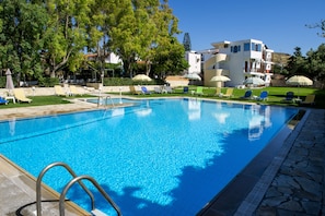 A clear blue swimming pool with lounge chairs and umbrellas, surrounded by trees and a modern building. - Prince Of Lillies (Hersonissos)
