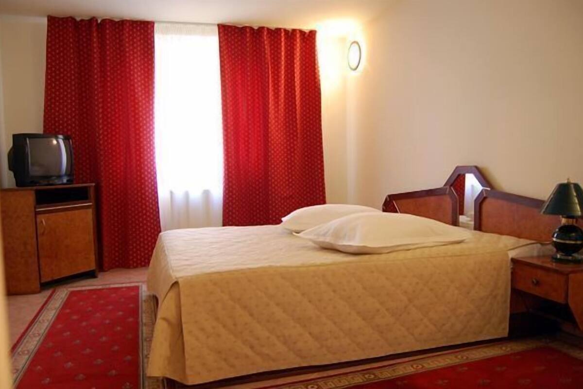 A hotel room with a bed, a television on a wooden cabinet, a lamp on a nightstand, and red curtains.