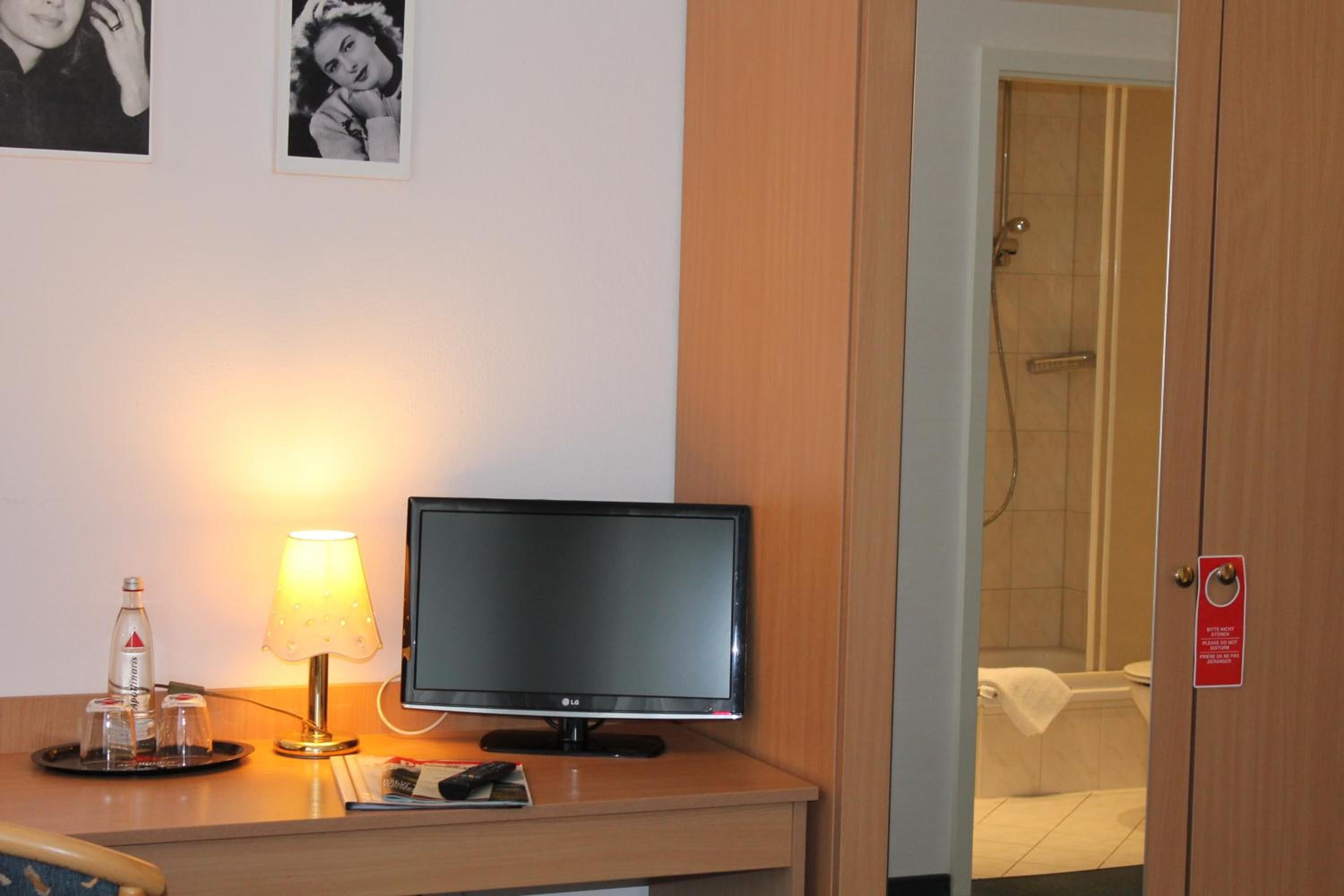 A hotel room with a desk, a flat-screen TV, a lamp, a bottle of water, and a towel rack.