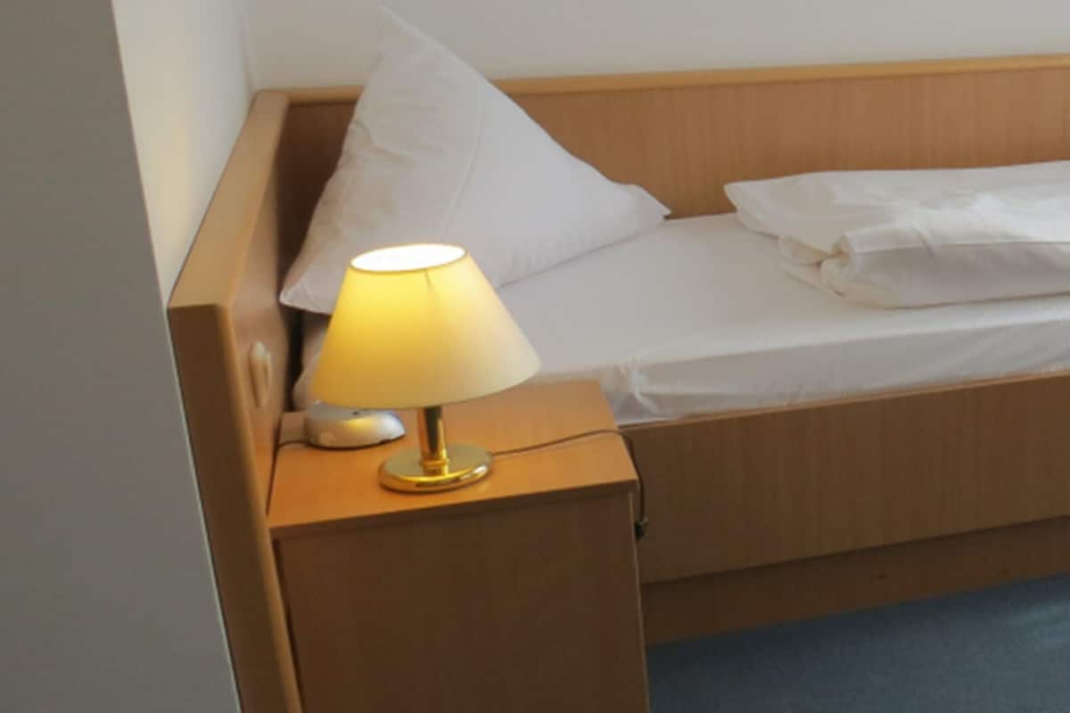 A single bed with white bedding, a bedside table with a lamp, and a window with blue curtains.