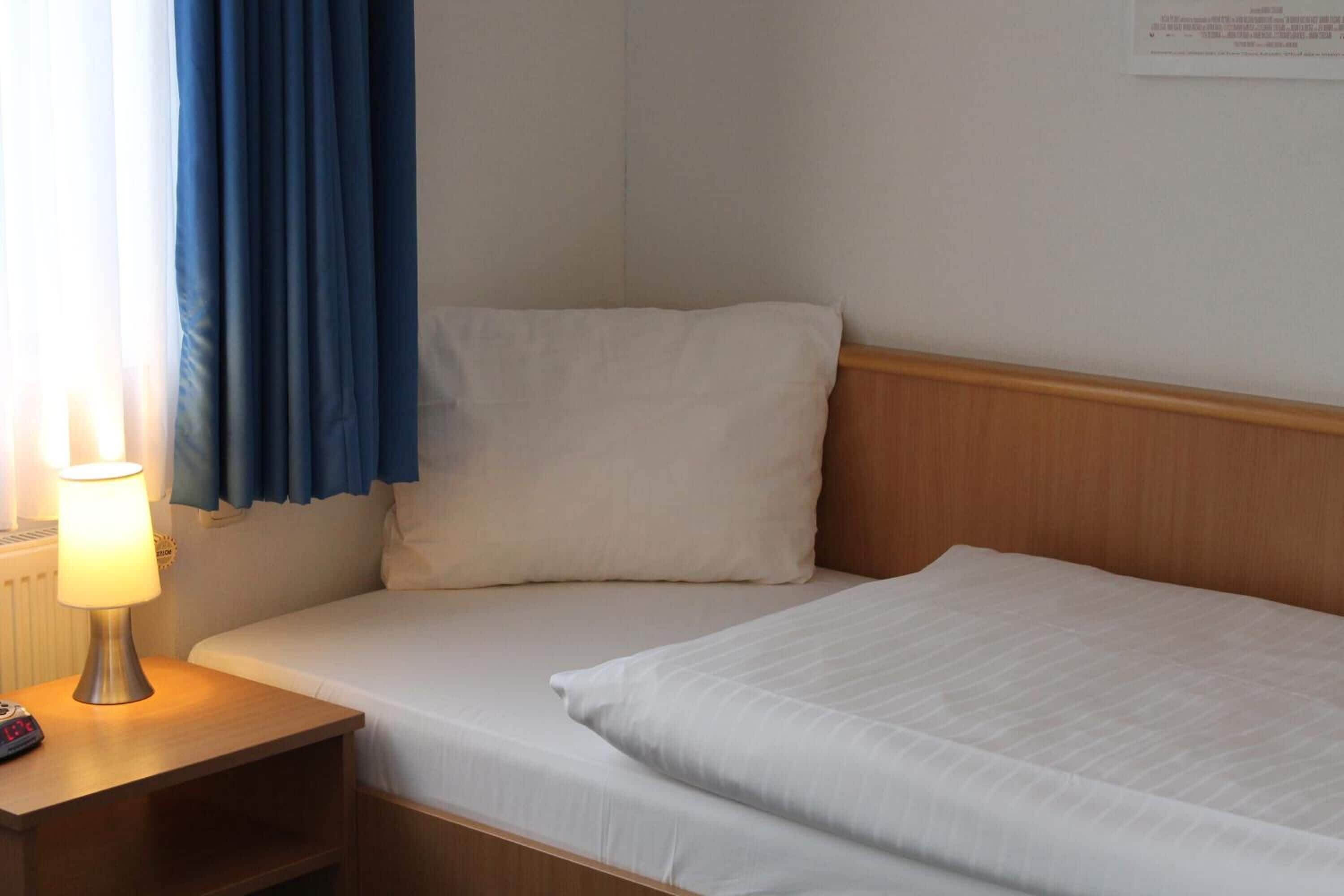 Un lit simple avec une literie blanche, une tête de lit en bois, une table de chevet avec une lampe et un rideau bleu.