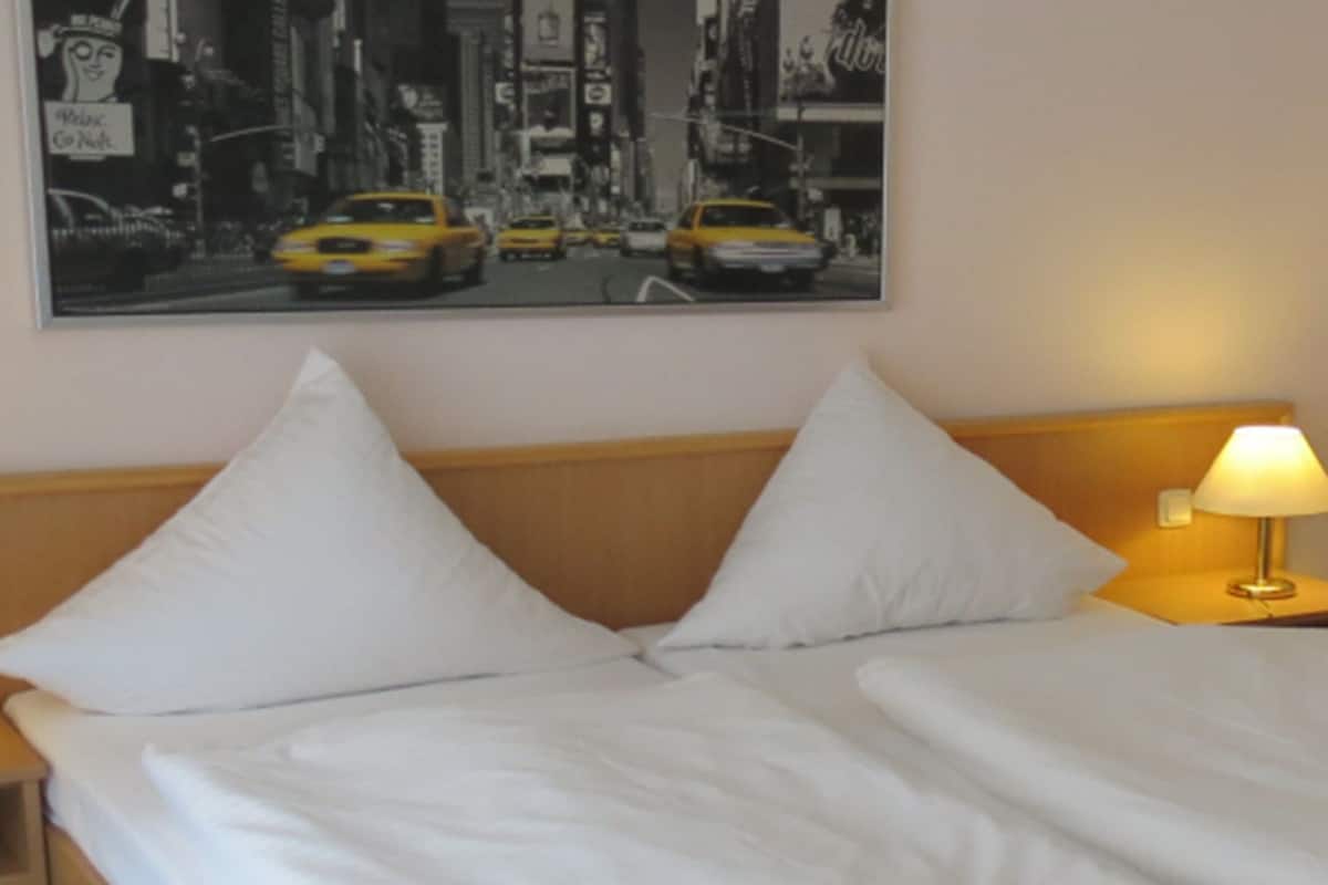 A hotel room with a bed, two bedside lamps, a nightstand with a phone, and a cityscape painting on the wall.
