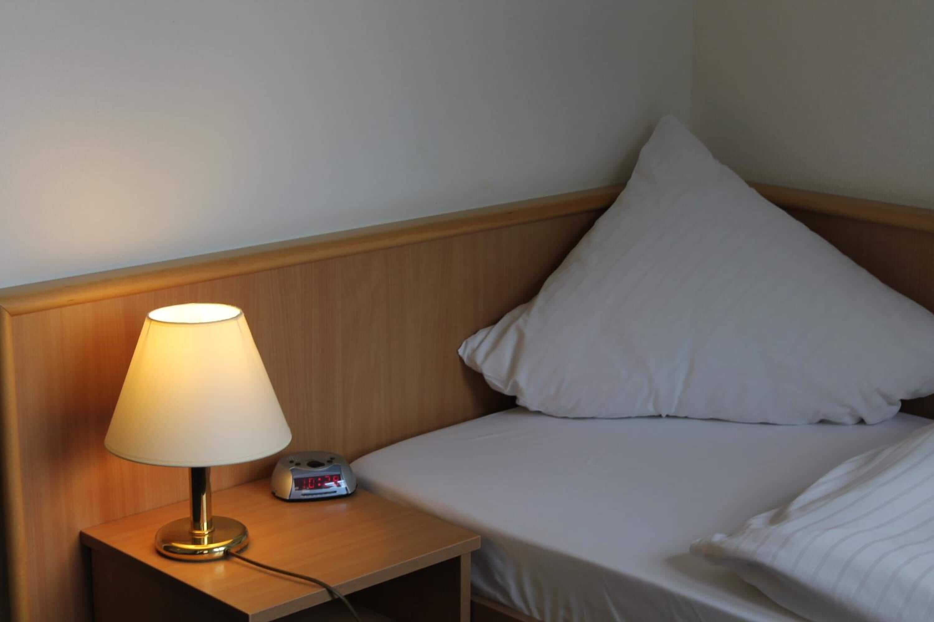 Una cama bien hecha con ropa blanca, un cabecero de madera, una mesita de noche con una lámpara y un reloj digital.