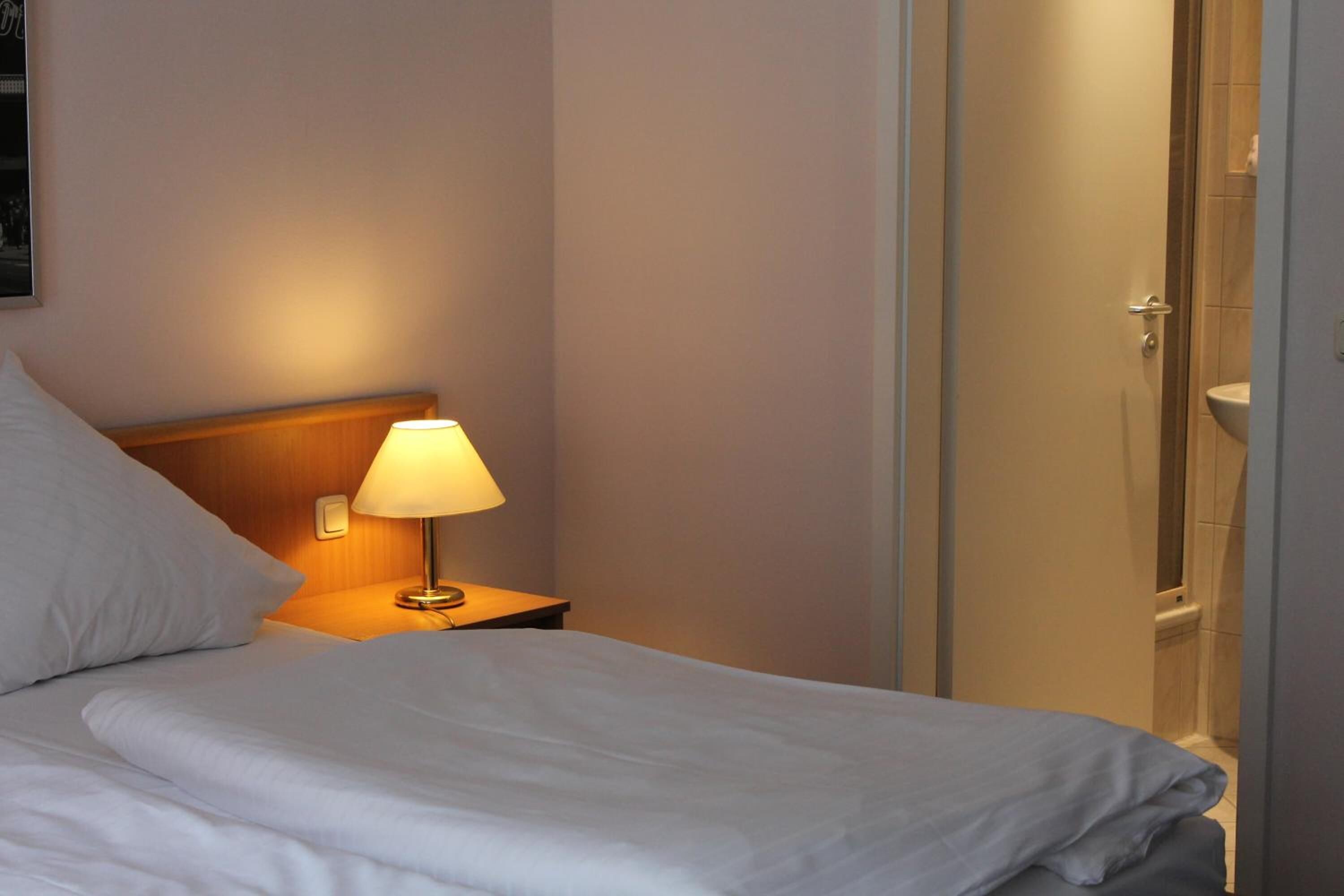 A neatly made bed with white linens, a bedside lamp, and a door leading to a bathroom.