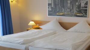 Habitación de hotel con una cama doble, dos mesitas de noche con lámparas, un cuadro grande con una vista urbana en la pared y una ventana con cortinas azules.