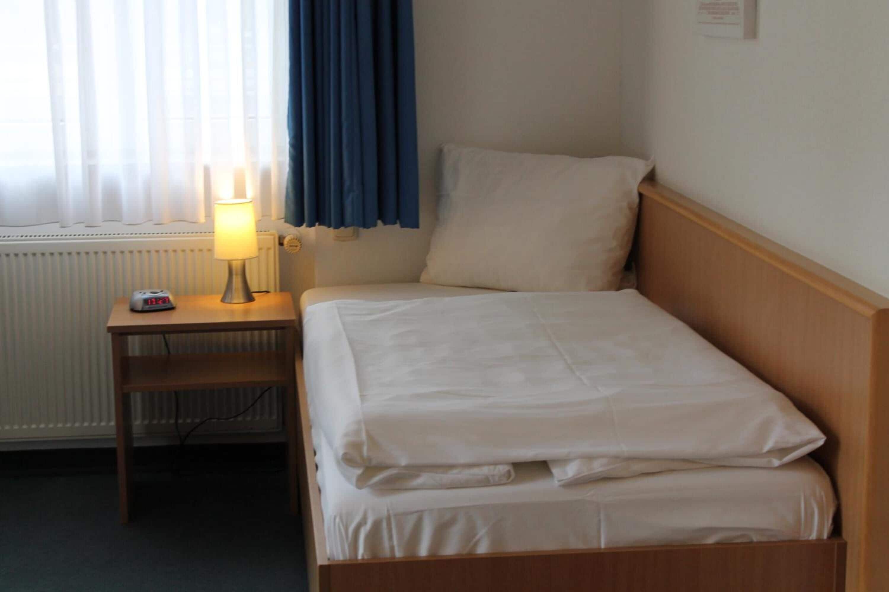 A single bed with white bedding, a bedside table with a lamp, and a window with curtains.