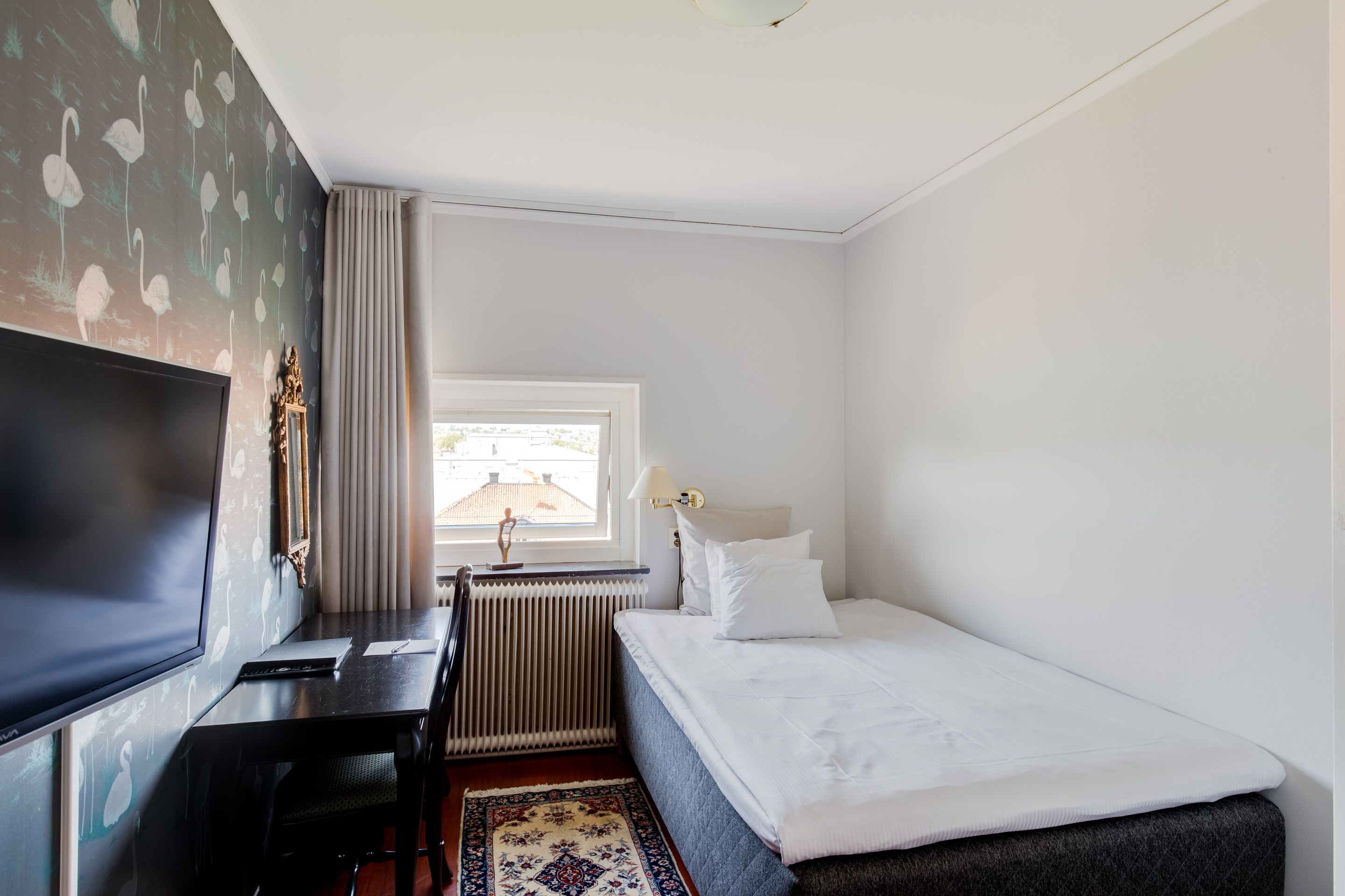 A hotel room with a bed, a desk, a television, a window with curtains, and a radiator.