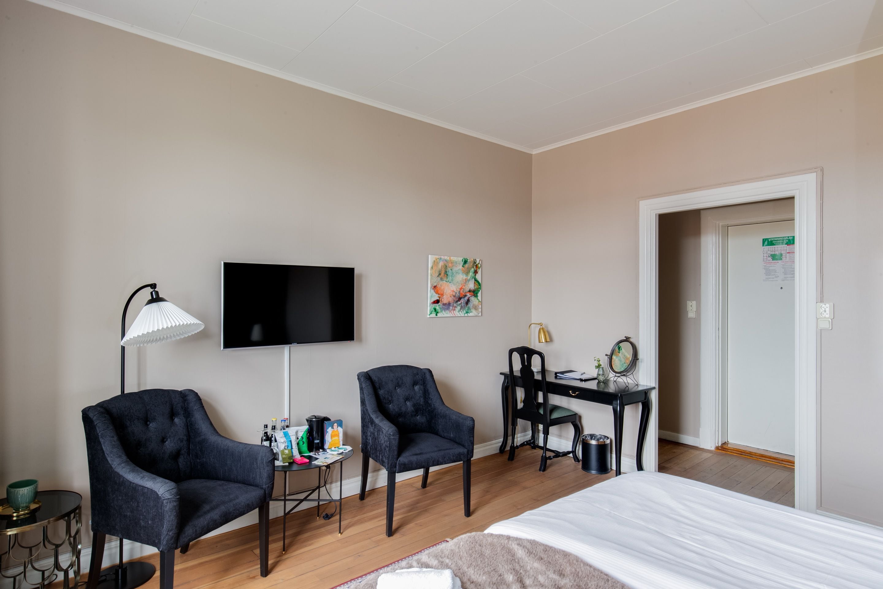 Ein Hotelzimmer mit einem Bett, zwei Sesseln, einem Schreibtisch mit Computer, einem Fernseher und einem kleinen Tisch mit Lampe.