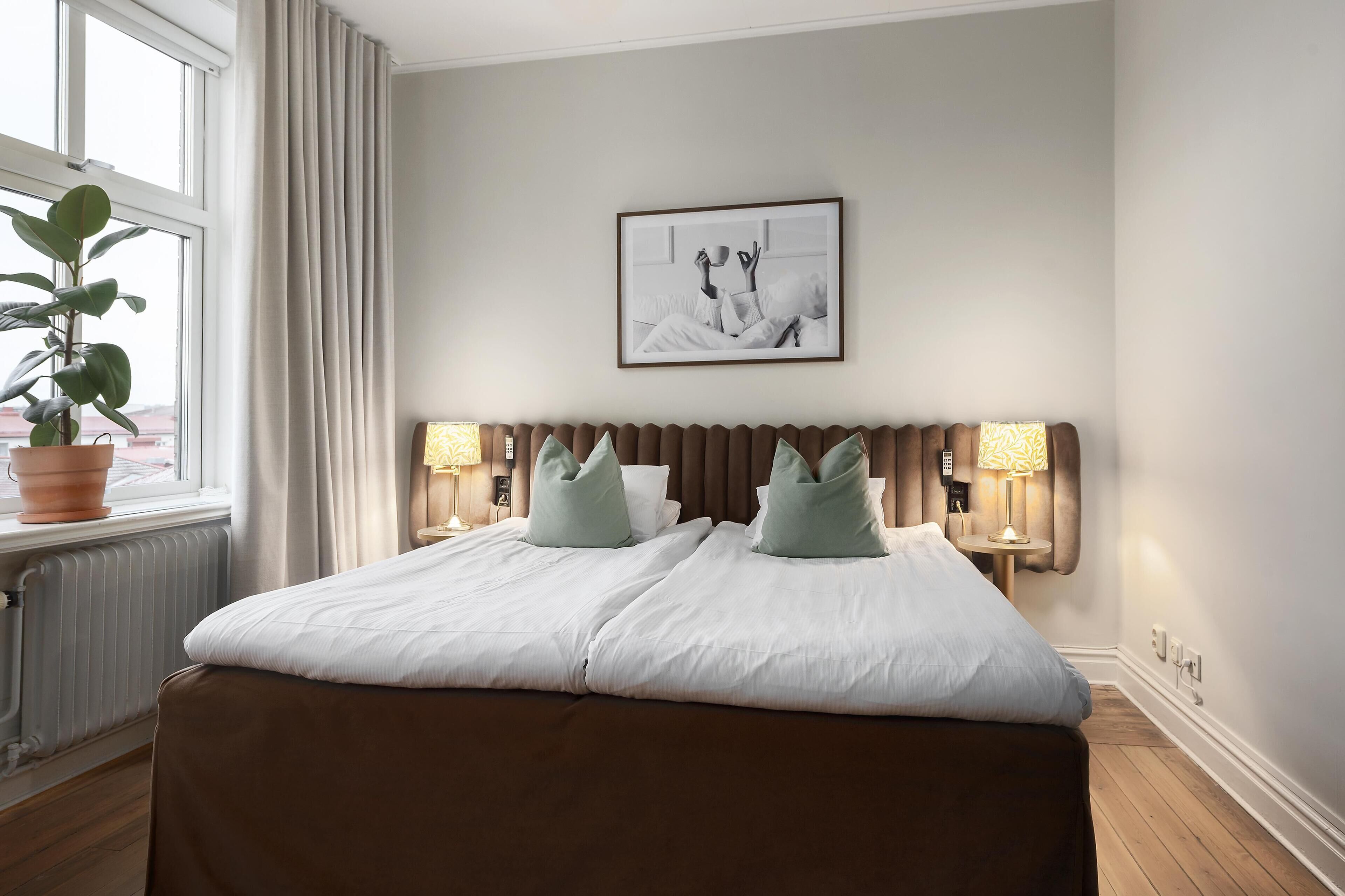 A hotel room with a large bed, two bedside tables with lamps, a radiator, a window with curtains, and a framed picture on the wall.