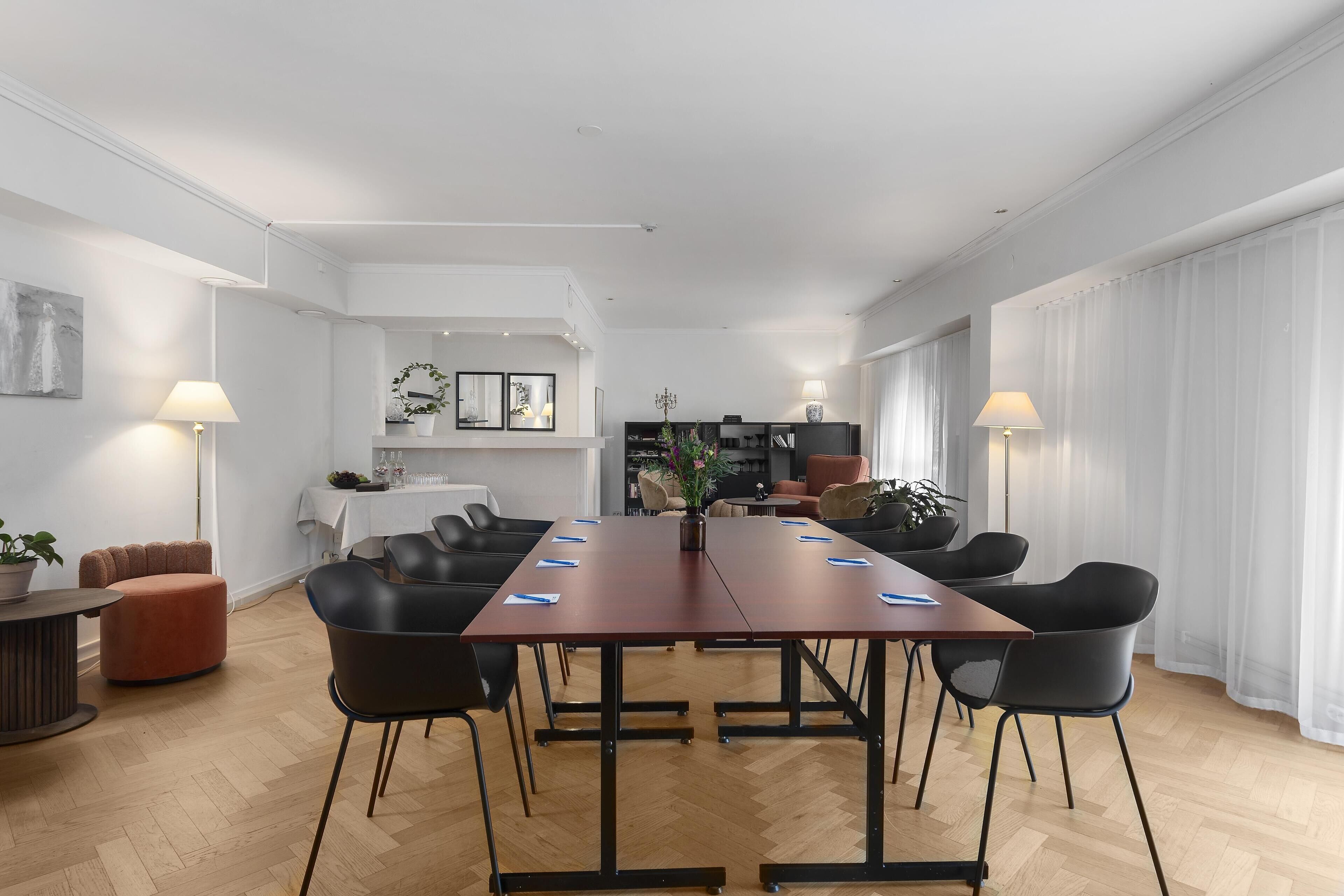 Una habitación amplia con una gran mesa de madera, sillas negras y una zona de comedor blanca al fondo.