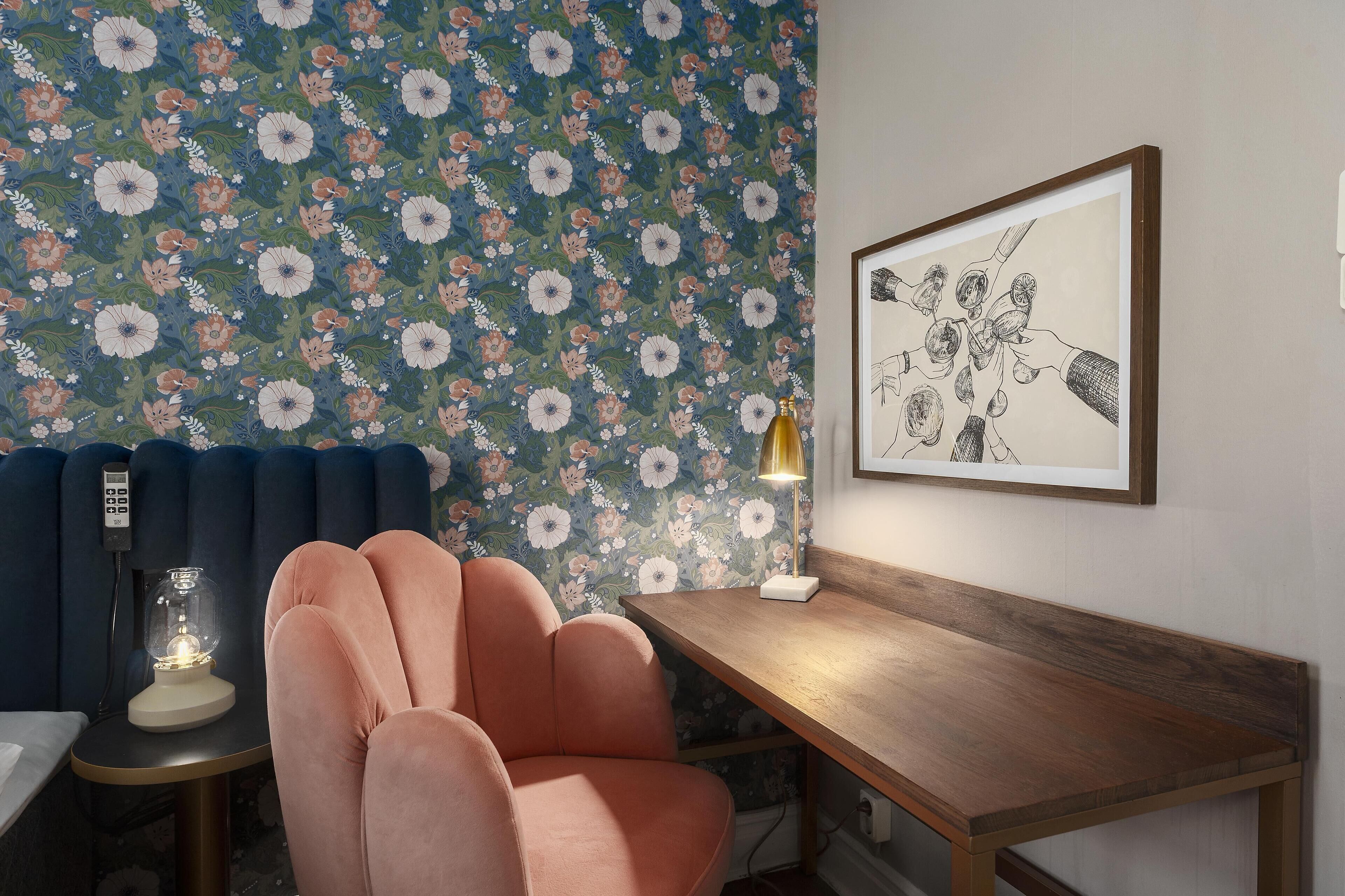 A room with a floral wallpaper, a wooden desk, a pink chair, and a framed artwork on the wall.