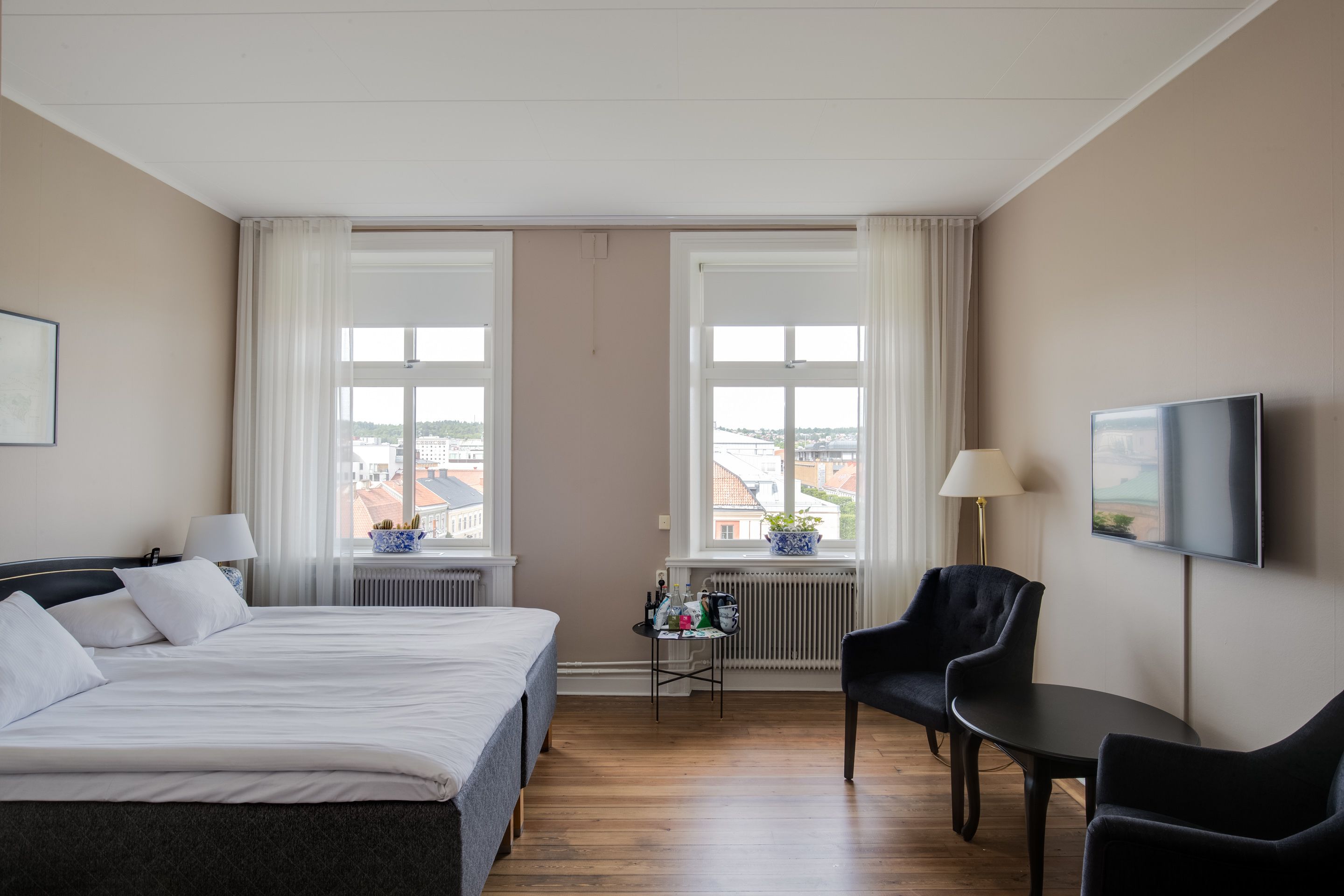 Hotelkamer met een bed, een bureau, twee stoelen, een televisie en uitzicht op gebouwen door grote ramen.