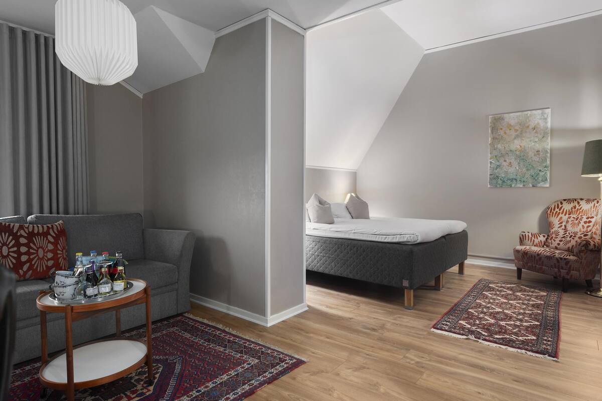 A modern living room with a grey sofa, a round wooden coffee table, a patterned rug, a bed with a grey duvet, and a chair with a floral pattern.