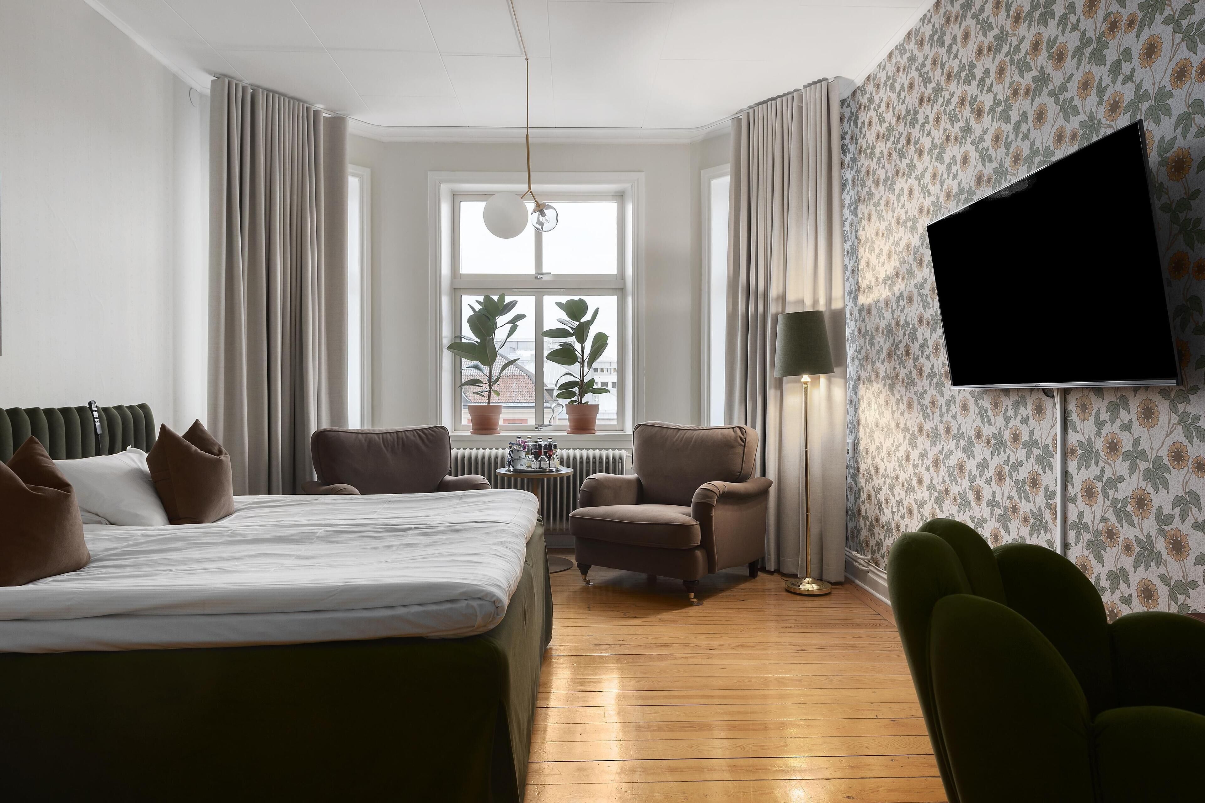 A hotel room with a bed, two armchairs, a small table, a TV, and a window with curtains.