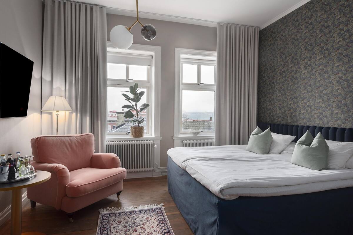 A bedroom with a bed, a chair, a table with bottles, and a view of the city.