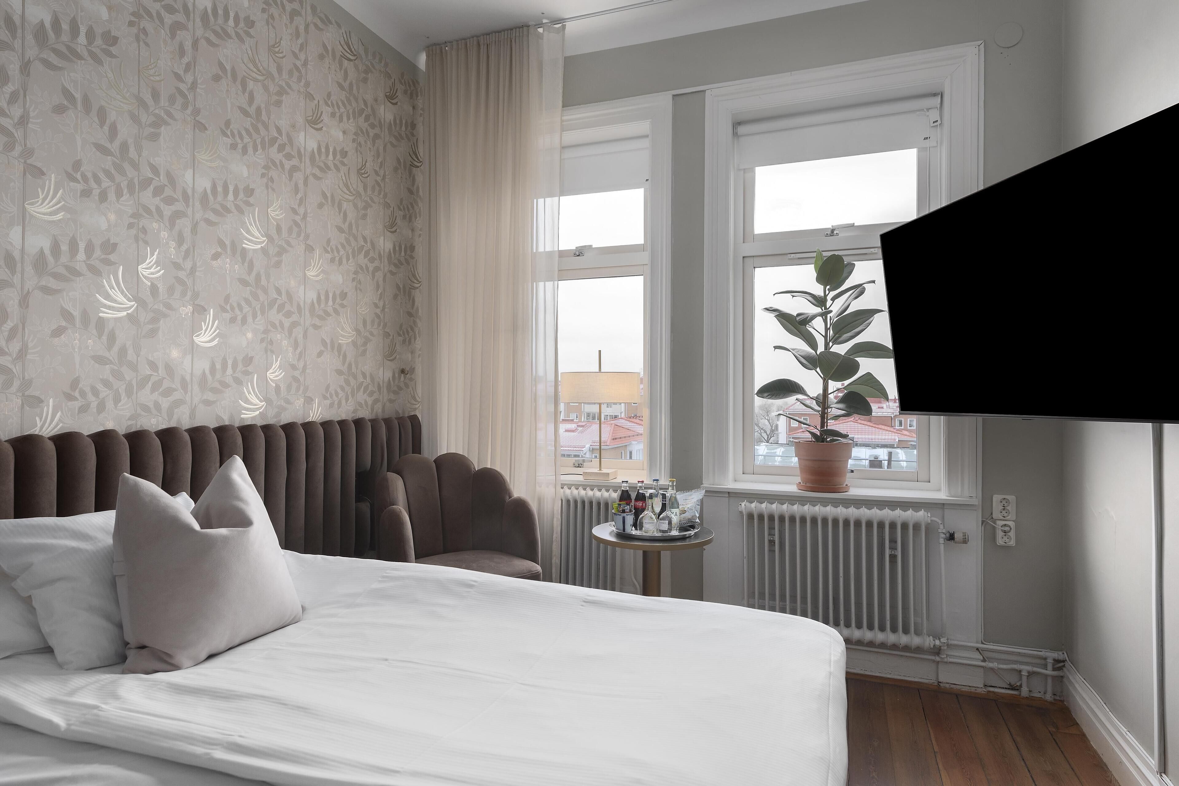 Et hotelværelse med seng, stol, lille bord med flasker, en potteplante og et vindue med udsigt til bygninger.