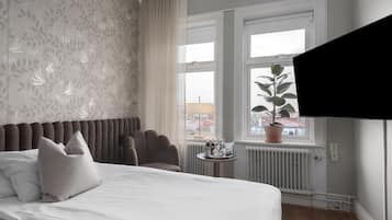 Ein Hotelzimmer mit einem Bett, einem Sessel, einem kleinen Tisch mit Flaschen, einer Topfpflanze und einem Fenster mit Blick auf Gebäude.