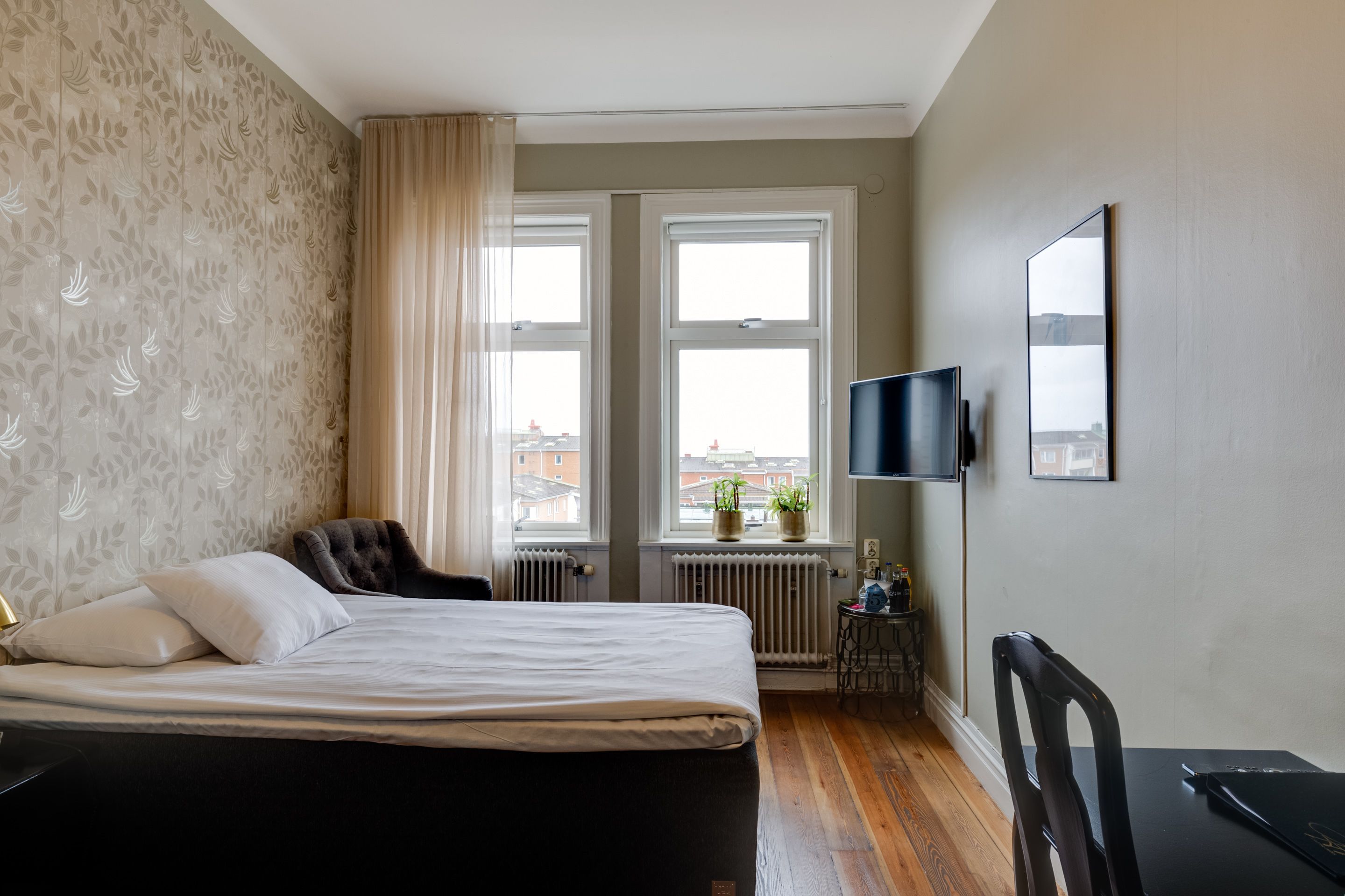 A bedroom with a bed, a TV, a chair, and a window with a view of buildings.