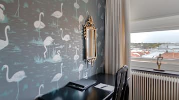 A room with a dark wall featuring a pattern of swans, a black table with a notebook and pen, a gold-framed mirror, and a view of buildings through a window.