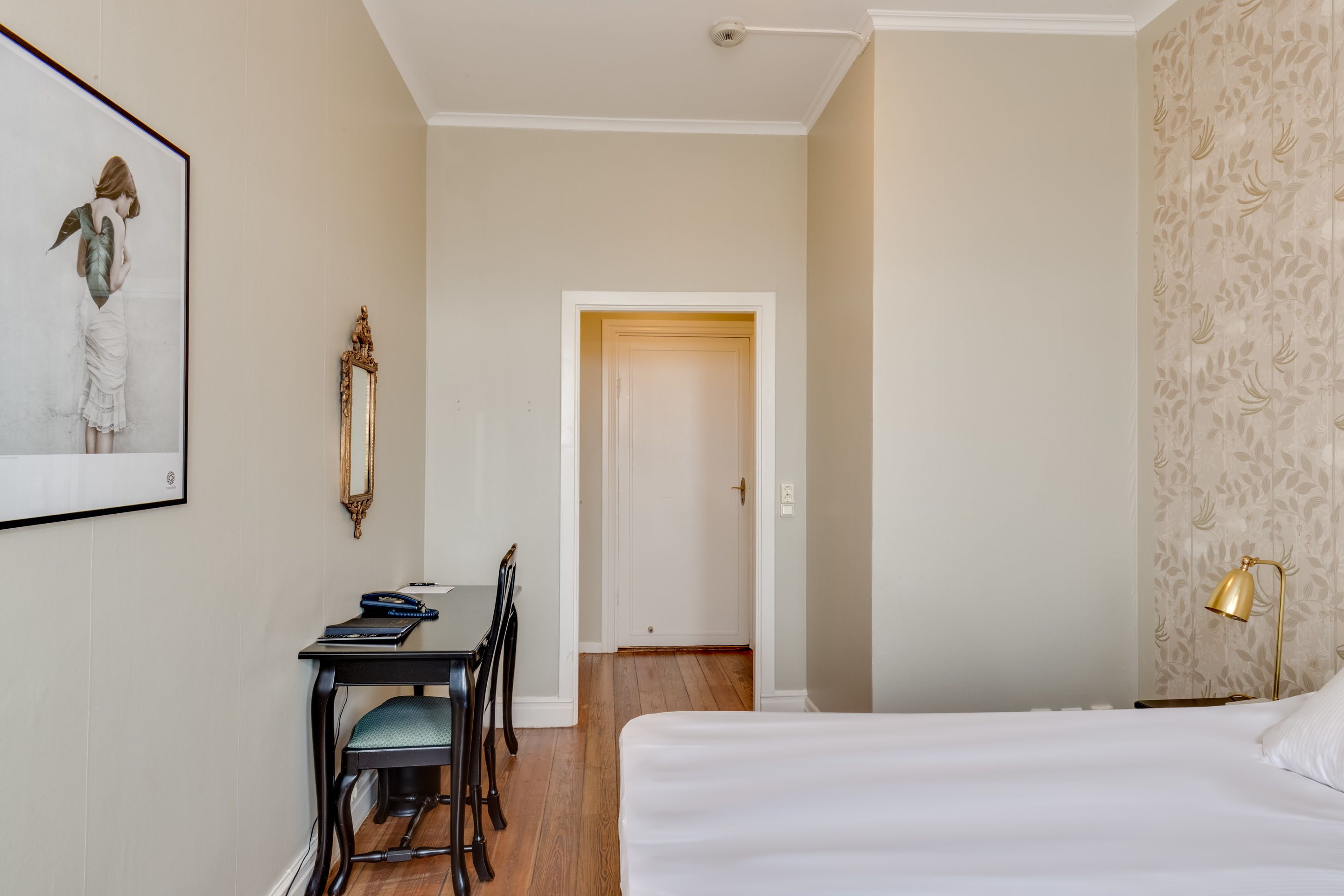 Ein Hotelzimmer mit einem Bett, einem Schreibtisch mit Stuhl, einem gerahmten Bild an der Wand und einer Tür zu einem weiteren Zimmer.