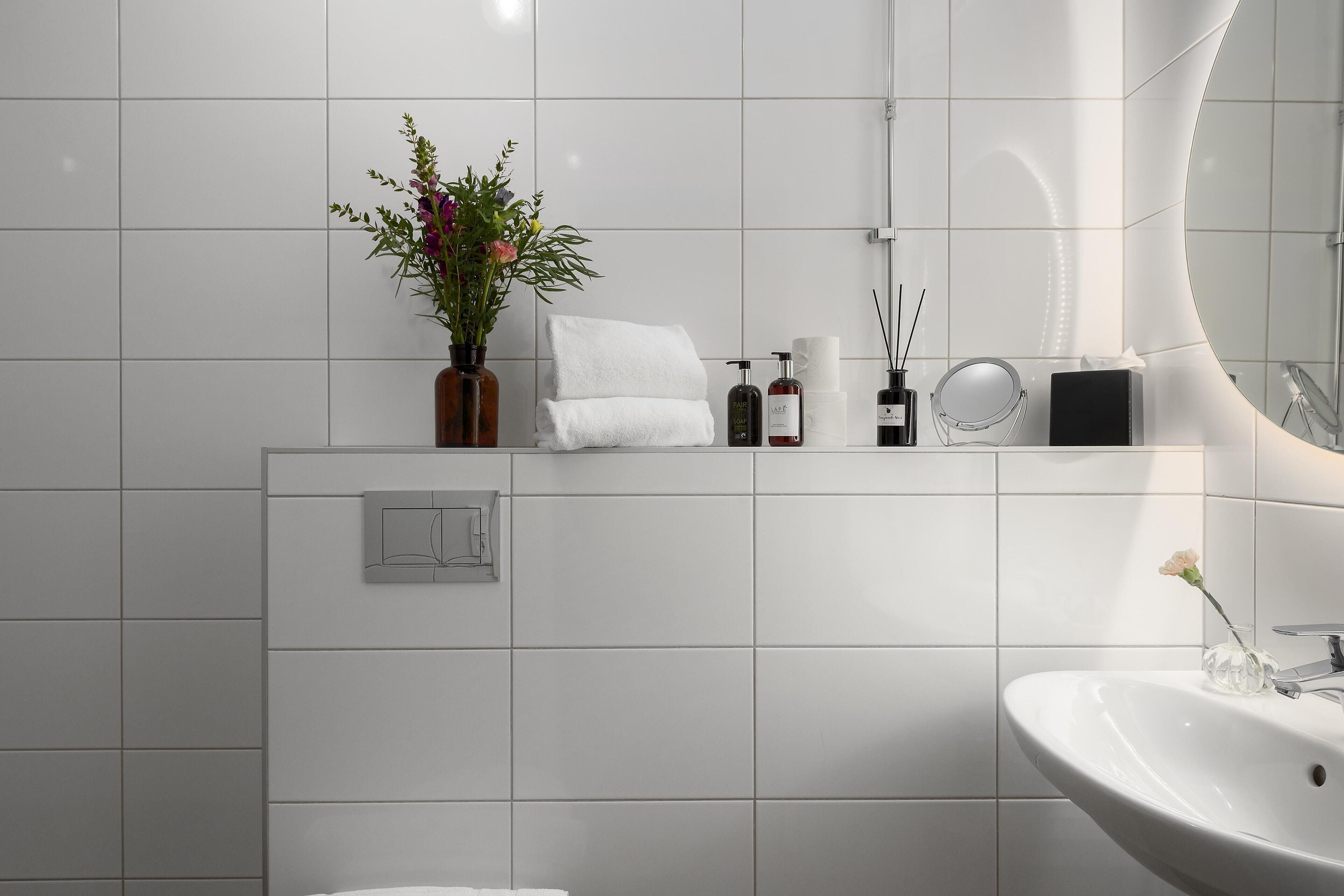 A modern bathroom with a white tiled wall, a large mirror, a sink, and a shelf with toiletries.