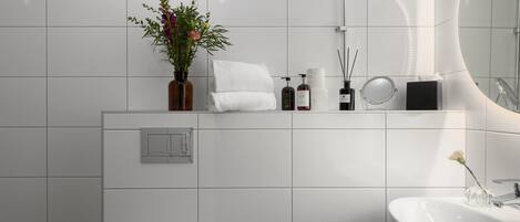 Un baño moderno con pared de azulejos blancos, un espejo grande, un lavabo y un estante con artículos de aseo.