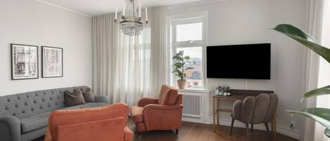 A living room with a grey sofa, orange armchairs, a wooden coffee table, a chandelier, a television, and a window with a view of buildings.