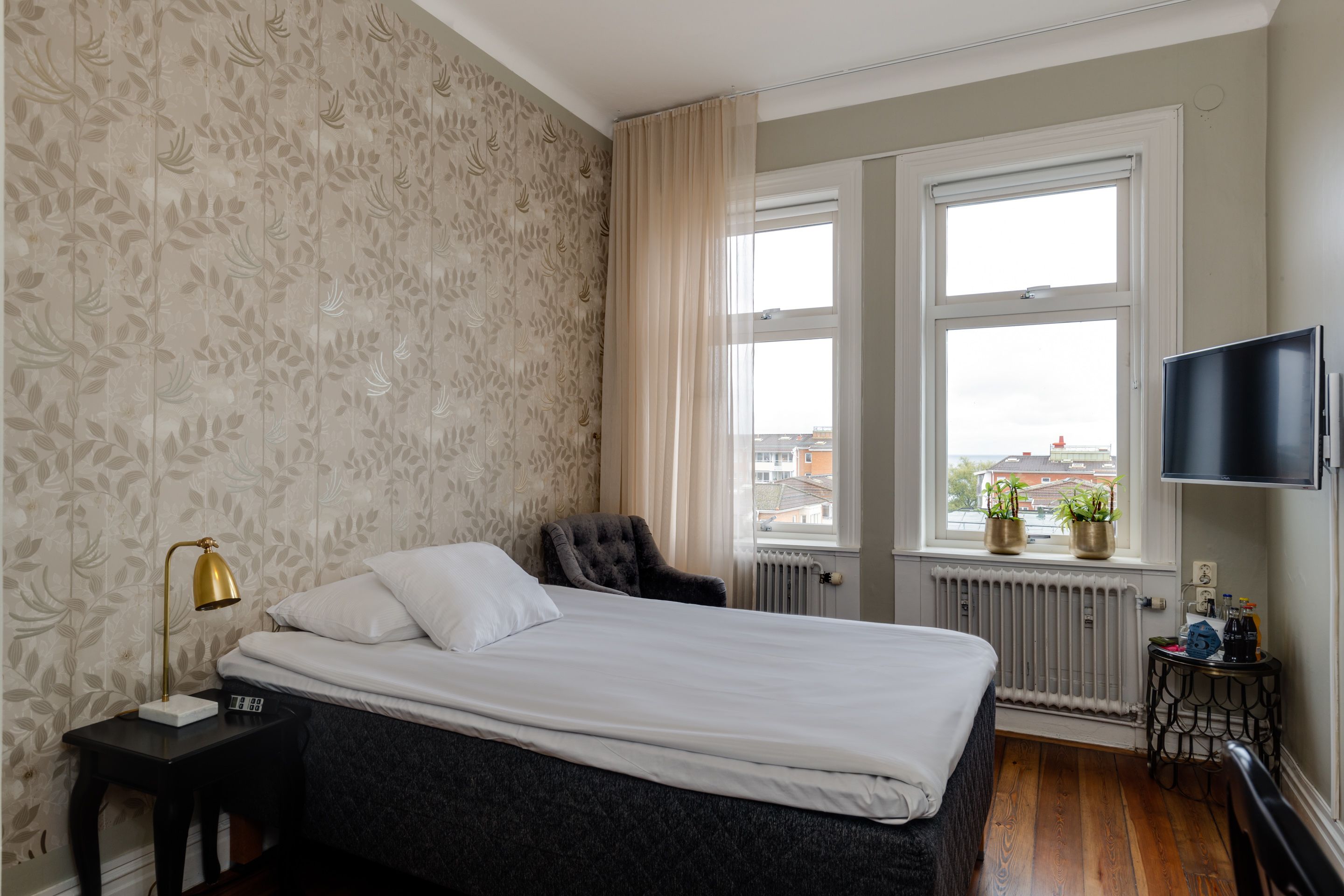 A bedroom with a bed, a nightstand, a chair, a window with curtains, a radiator, and a TV mounted on the wall.
