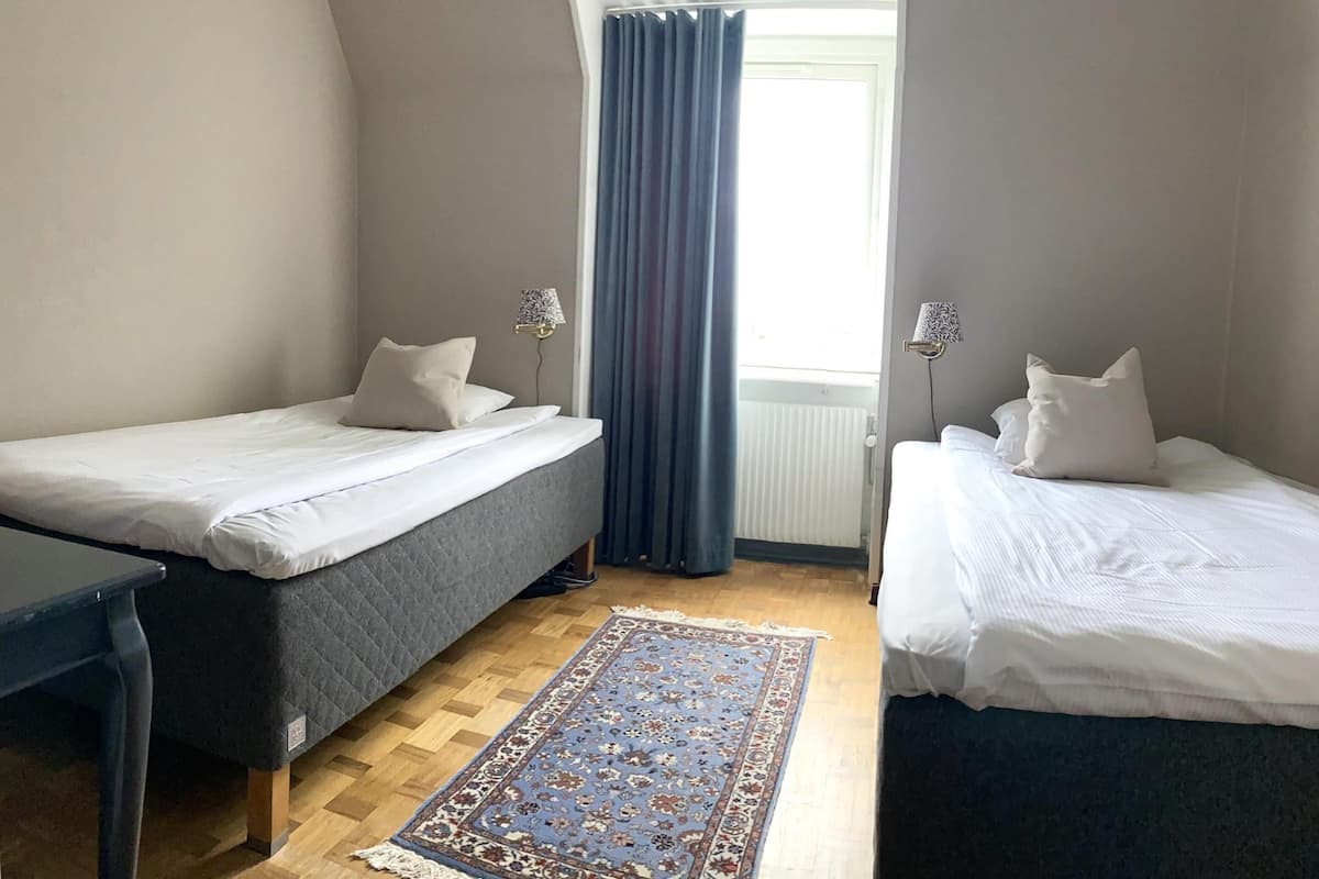 A room with two beds, a wooden floor, a patterned rug, and a window with curtains.