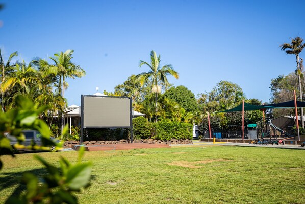 Un espacio con césped, una pantalla grande en blanco, palmeras y un parque infantil.