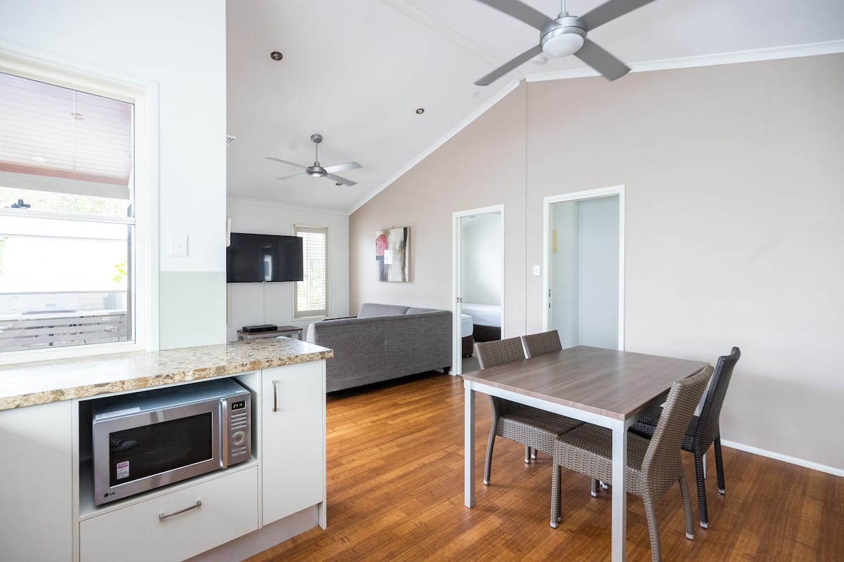 Une cuisine compacte équipée d’un four à micro-ondes, d’une table à manger avec des chaises, d’un ventilateur de plafond et d’un canapé gris, le tout dans un appartement moderne.