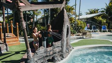 Una estructura de juego en forma de barco pirata de madera sobre una piscina, rodeada de palmeras y sillas de descanso.
