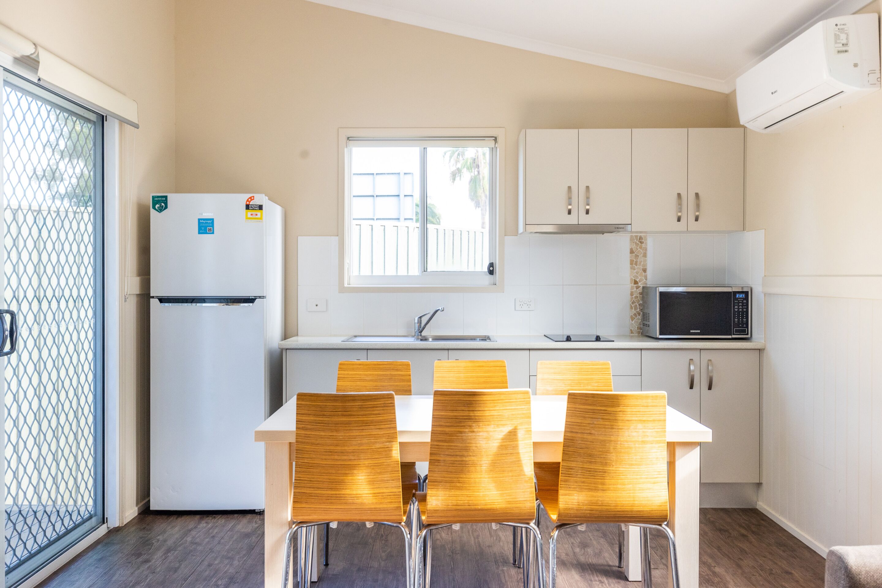 A compact kitchen with a refrigerator, microwave, and white cabinets. There is a dining table with chairs and a window with a view.