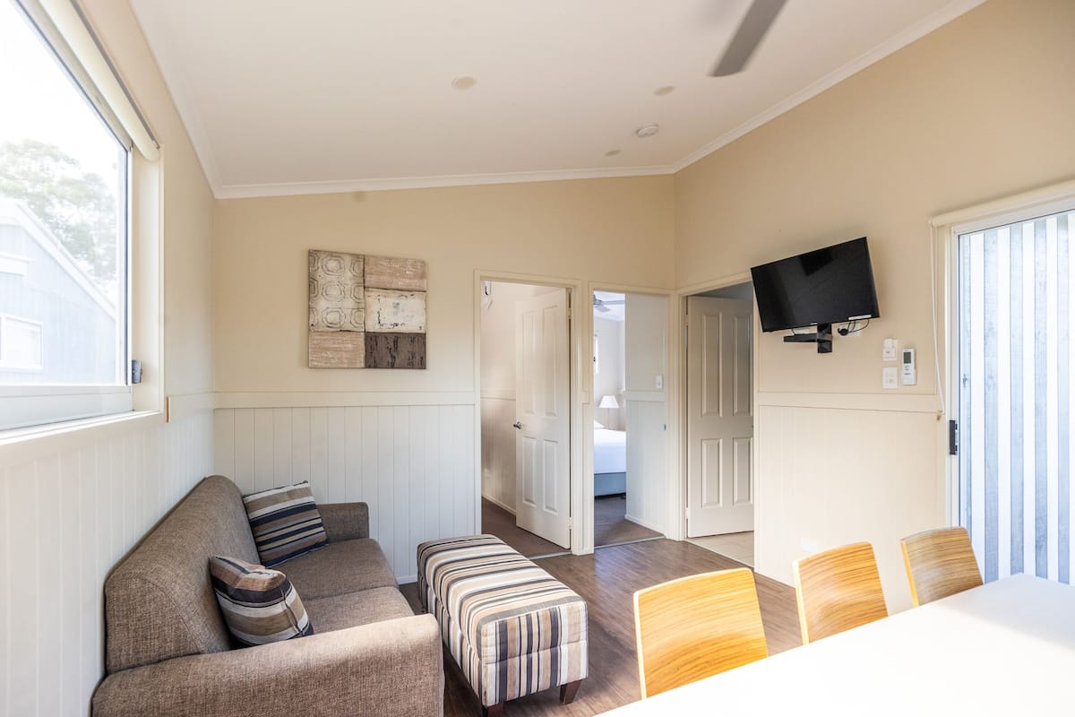 Un salon comprenant un canapé, un pouf et une table à manger avec des chaises. Un téléviseur est fixé au mur et une porte donne accès à une autre pièce.
