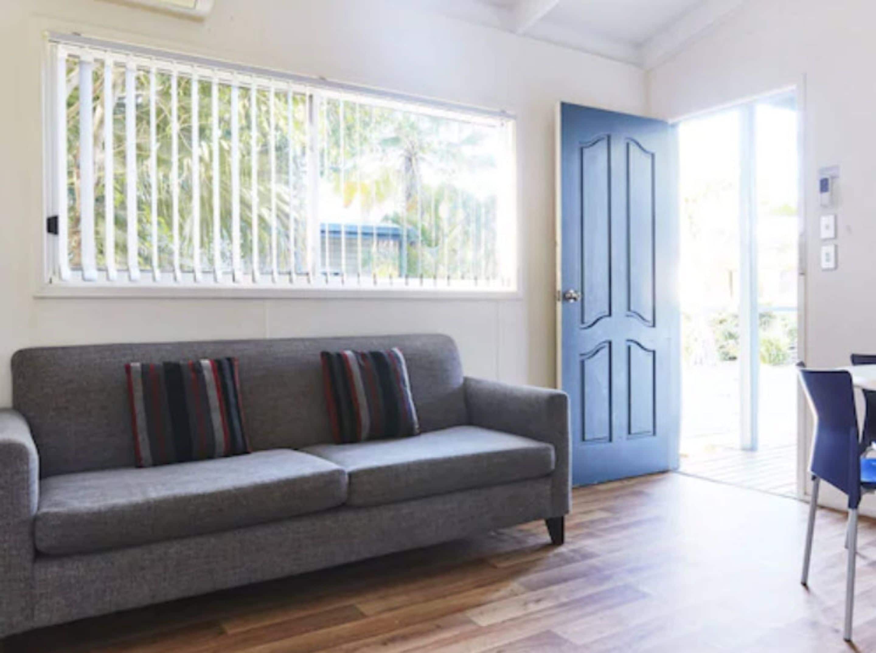 Una habitación con un sofá gris, una puerta azul y una ventana con persianas.