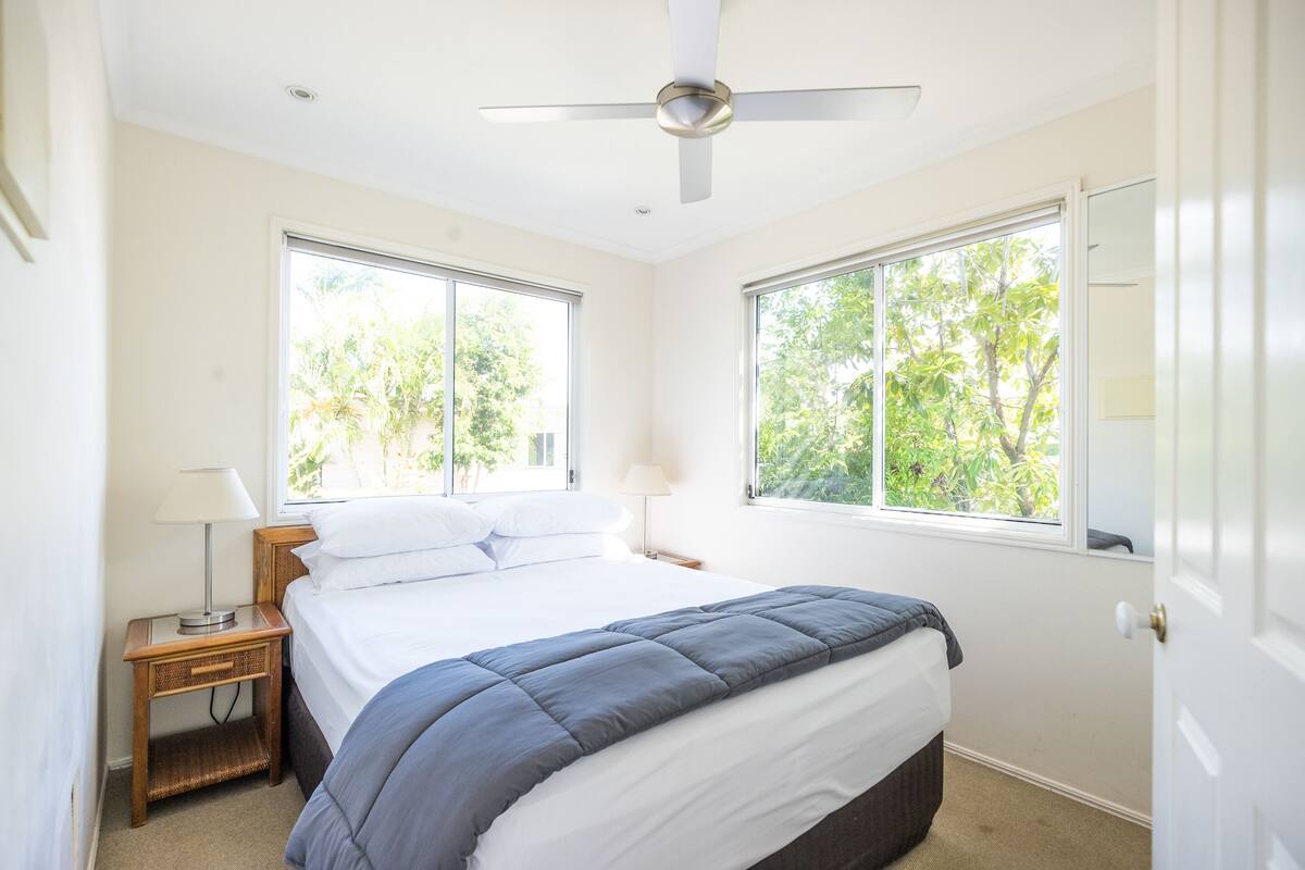 Une chambre à coucher comprenant un lit, deux tables de chevet, un ventilateur de plafond et de grandes fenêtres donnant sur un paysage verdoyant.