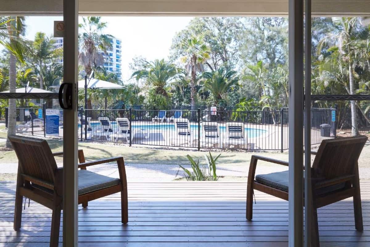 Un espace aménagé au bord de la piscine, avec deux chaises en bois, une piscine et une vue sur des palmiers et des bâtiments.