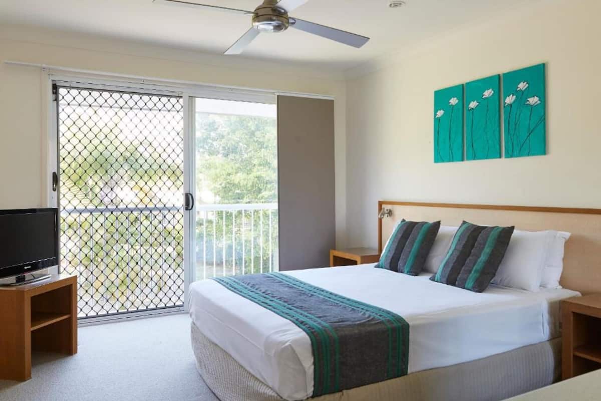 Une chambre avec un lit, une télévision, un ventilateur de plafond et un balcon donnant sur un paysage verdoyant.