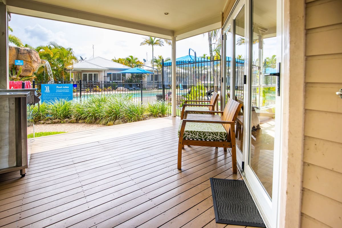 Une terrasse en bois avec une porte vitrée donnant sur une zone de piscine, des meubles d’extérieur et un panneau indiquant les règles à respecter au bord de la piscine.