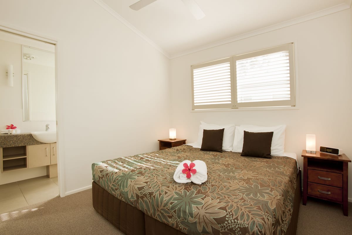 Une chambre d’hôtel avec un grand lit, une table de chevet, une commode, un miroir, une fenêtre avec des stores et une salle de bain équipée d’un lavabo et d’un miroir.