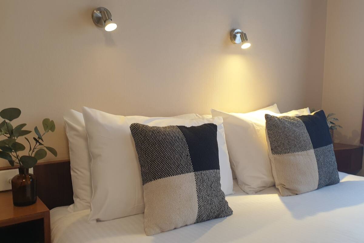 Ein ordentlich bezogenes Bett mit weißer Bettwäsche und zwei Zierkissen, ein Nachttisch mit Pflanze sowie Wandleuchten.