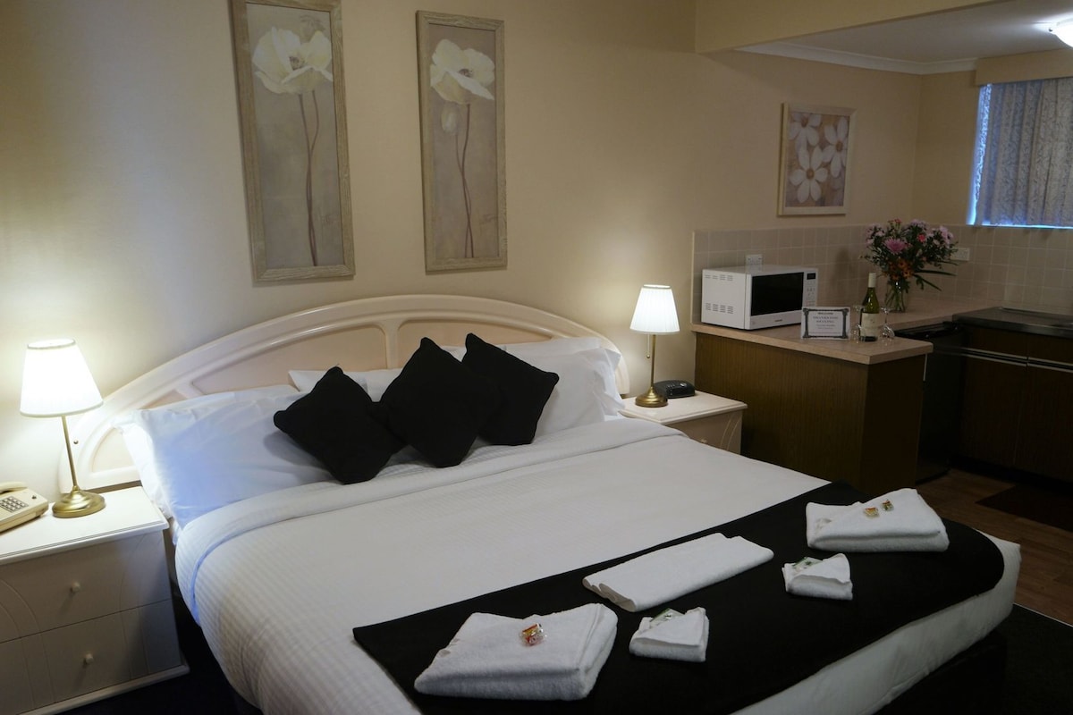 A hotel room with a bed, bedside lamps, a microwave, a small table with a vase of flowers, and artwork on the walls.