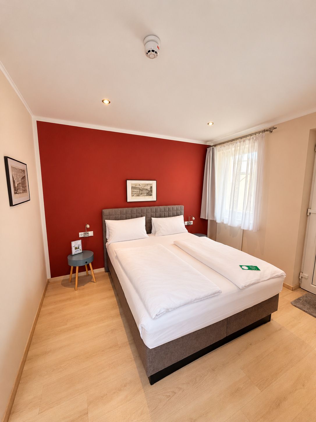 Ein Hotelzimmer mit einem Bett, einem kleinen Nachttisch, einem gerahmten Bild an der Wand und einem Fenster mit Vorhängen.