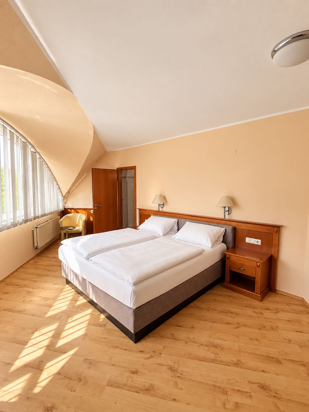 A bedroom with a large bed, two bedside tables, a chair, and a window with blinds.