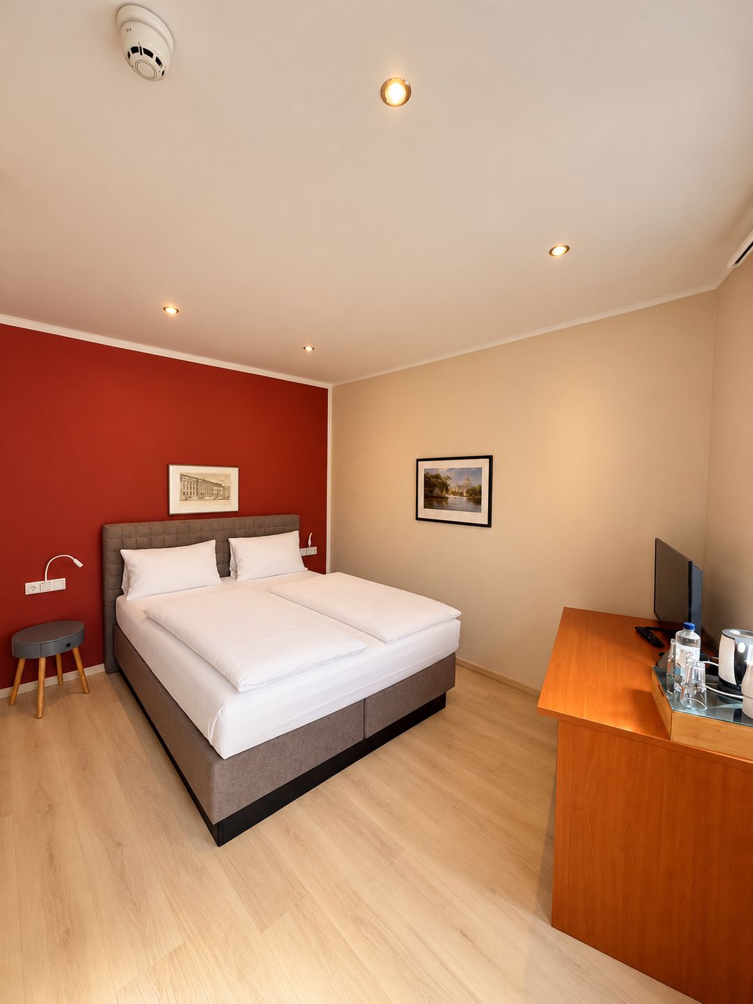A hotel room with a bed, a desk, a chair, a small side table, and a wall-mounted TV.
