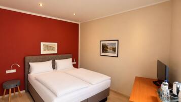 A hotel room with a bed, a desk, a chair, a small side table, and a wall-mounted TV.