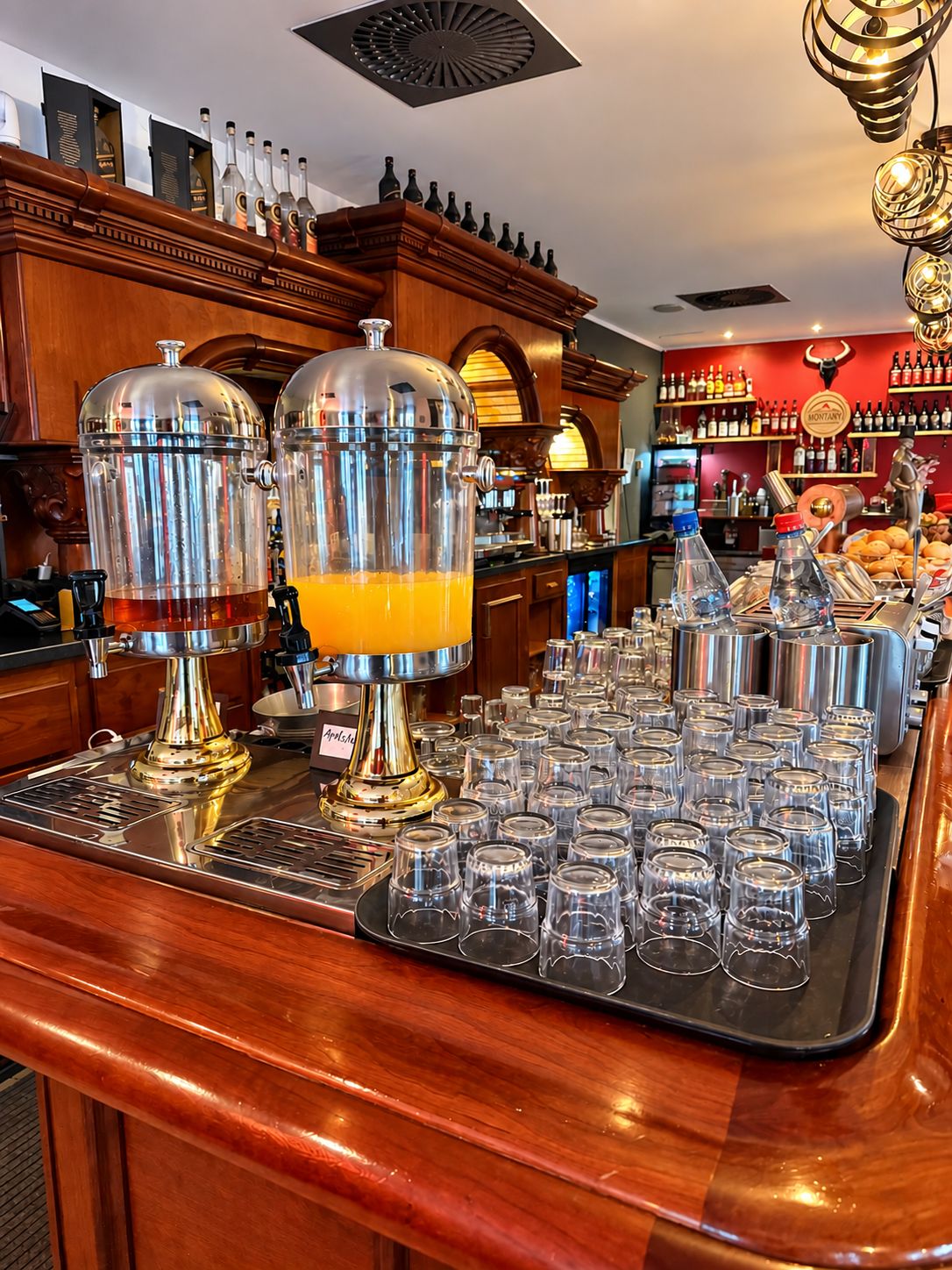 A bar area with two large beverage dispensers, a tray of glasses, and a variety of bottles in the background.
