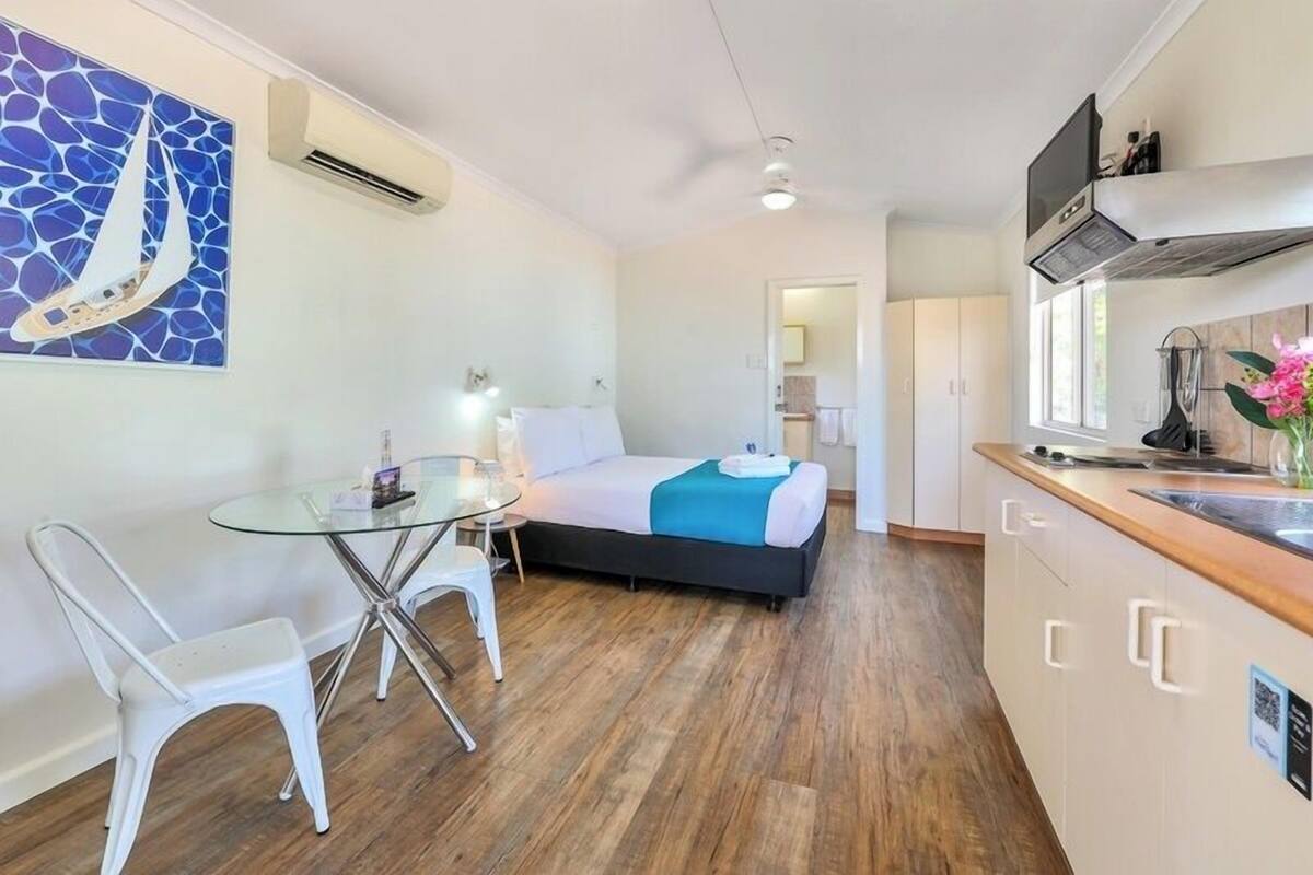 Habitación de hotel compacta con cama, una mesita pequeña, una silla, una mesita de vidrio, kitchenette y un baño visible a través de una puerta abierta.