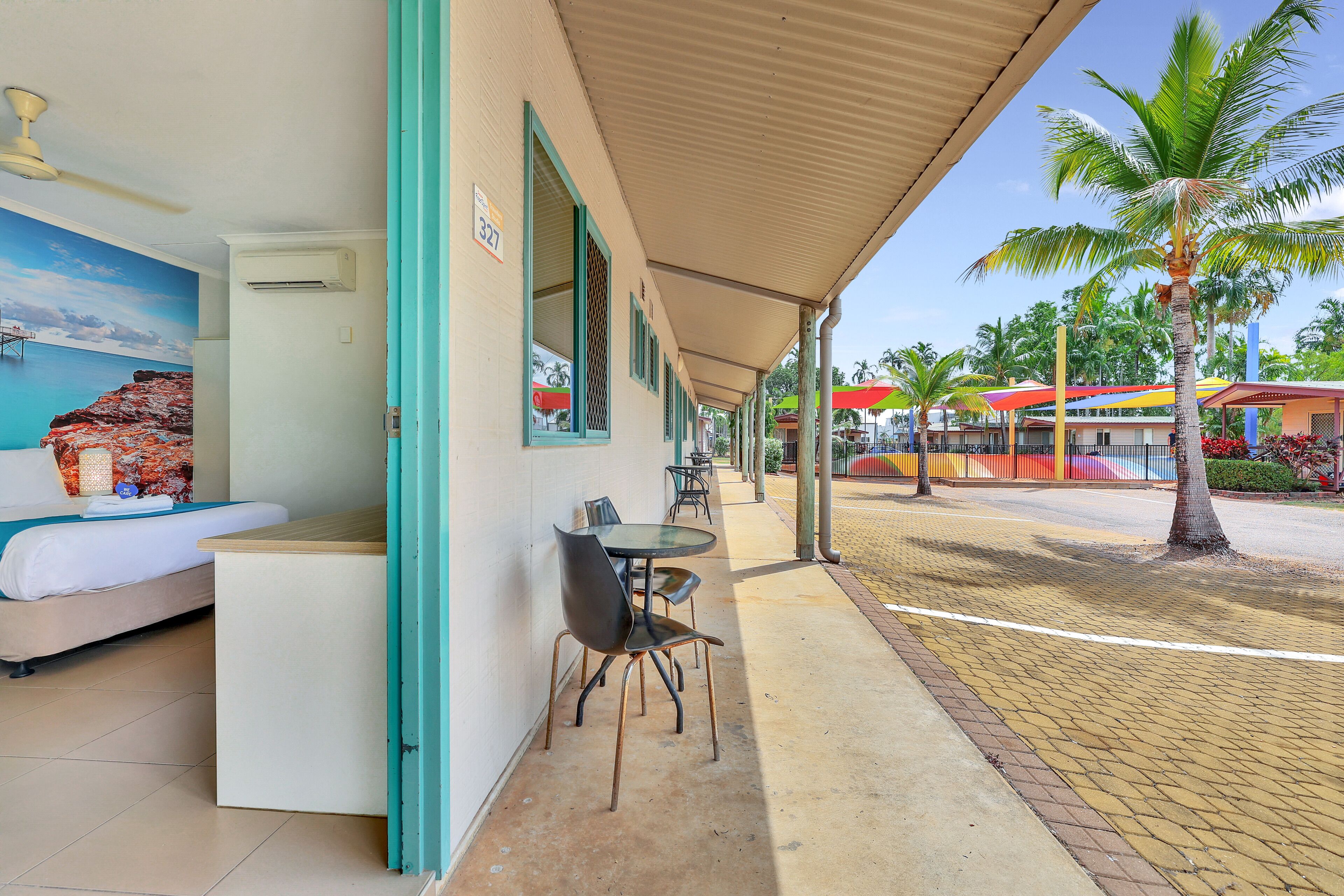 A hotel room with a bed, a small table, and a chair. There is a balcony with a view of the street and palm trees.