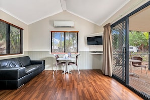 A room with a black leather sofa, a dining table with four chairs, a flat-screen TV mounted on the wall, and a sliding glass door leading to an outdoor area.